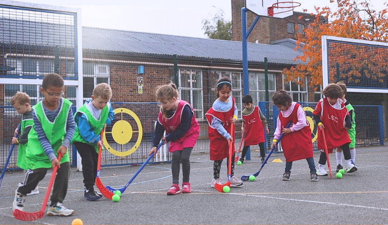 Children (a mix of boys and girls), standing in a line wearing colourful bibs and holding Hockey sticks on a school playground. This photo was taken at an LSC holiday club.