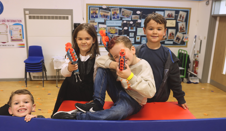 3 children holding Nerf equipment with smiles, preparing for Nerf Adventures at LSC Holiday Club. Child at the bottom of the screen, poking out from play equipment smiling