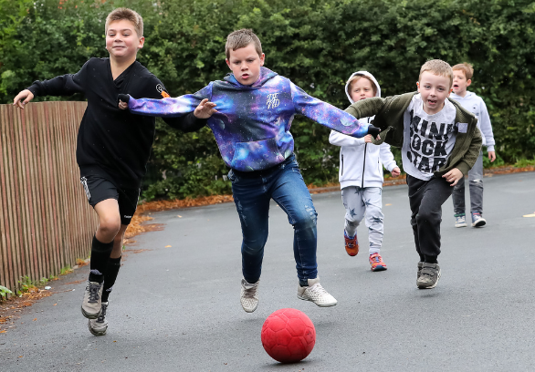 Three children (boys) running towards the camera in a game of football on a primary school playground. They are running towards a red foam football